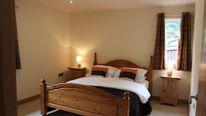A bedroom with a wooden bed, two bedside tables with lamps, a window with curtains, and a door. - Loch Ness Highland Cottages (Inverness)