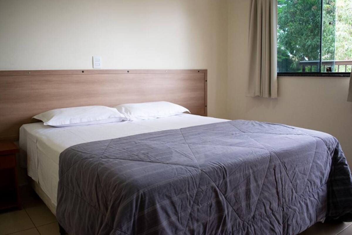 A bed with a wooden headboard, white bedding, and a view of greenery through a window.