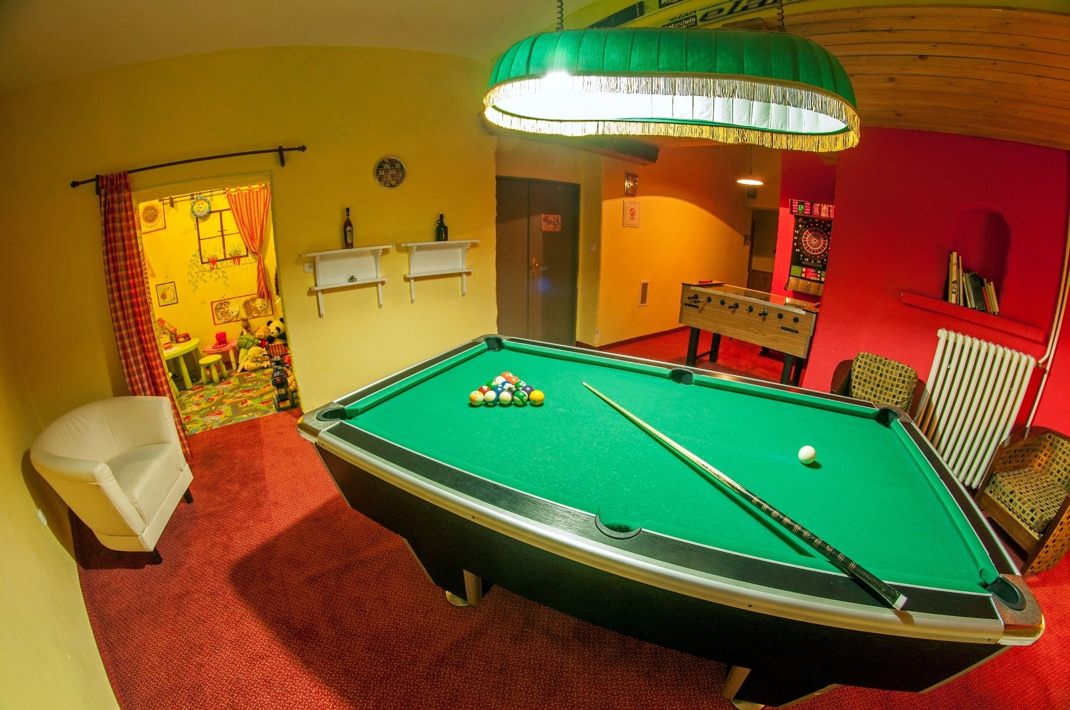 Ein Billardtisch mit Kugeln, ein Sofa, eine Lampe und ein Kicker in einem Raum mit roten Wänden und Teppichboden.
