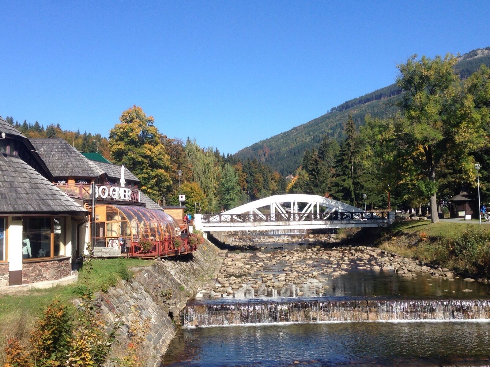 A rustic building with a glass greenhouse, a wooden bridge, and a river.