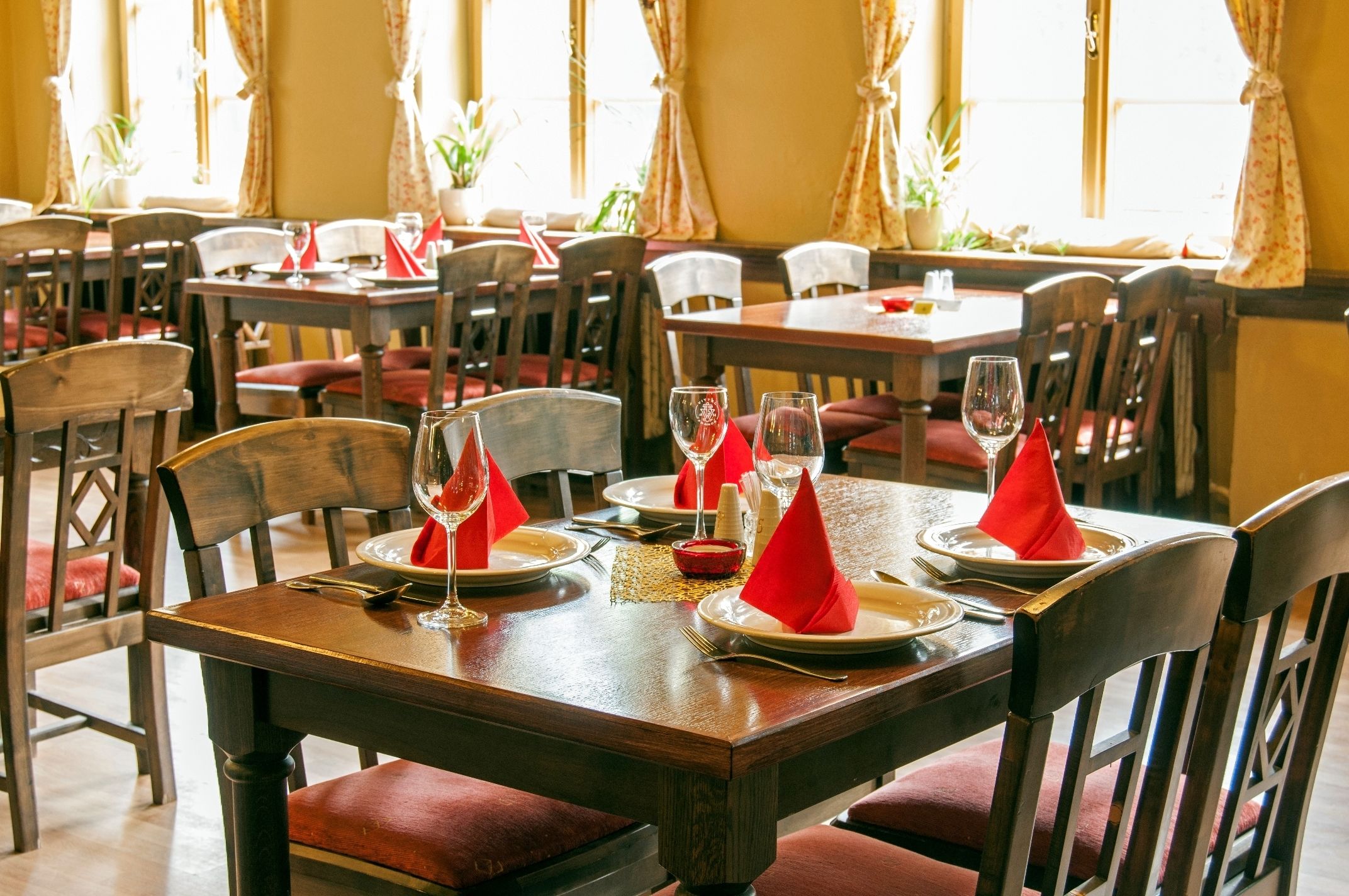 A dining area with wooden tables and chairs, red napkins, and wine glasses set for a meal.