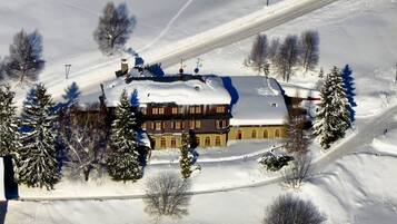 Un edificio grande de varios pisos con techo nevado, rodeado de árboles cubiertos de nieve y una pista de esquí.