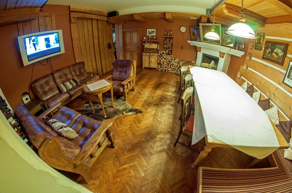 A cozy living room with a wooden herringbone floor, leather sofas, a dining table with a white tablecloth, and a television mounted on the wall. - Alpský hotel (Spindleruv Mlyn)