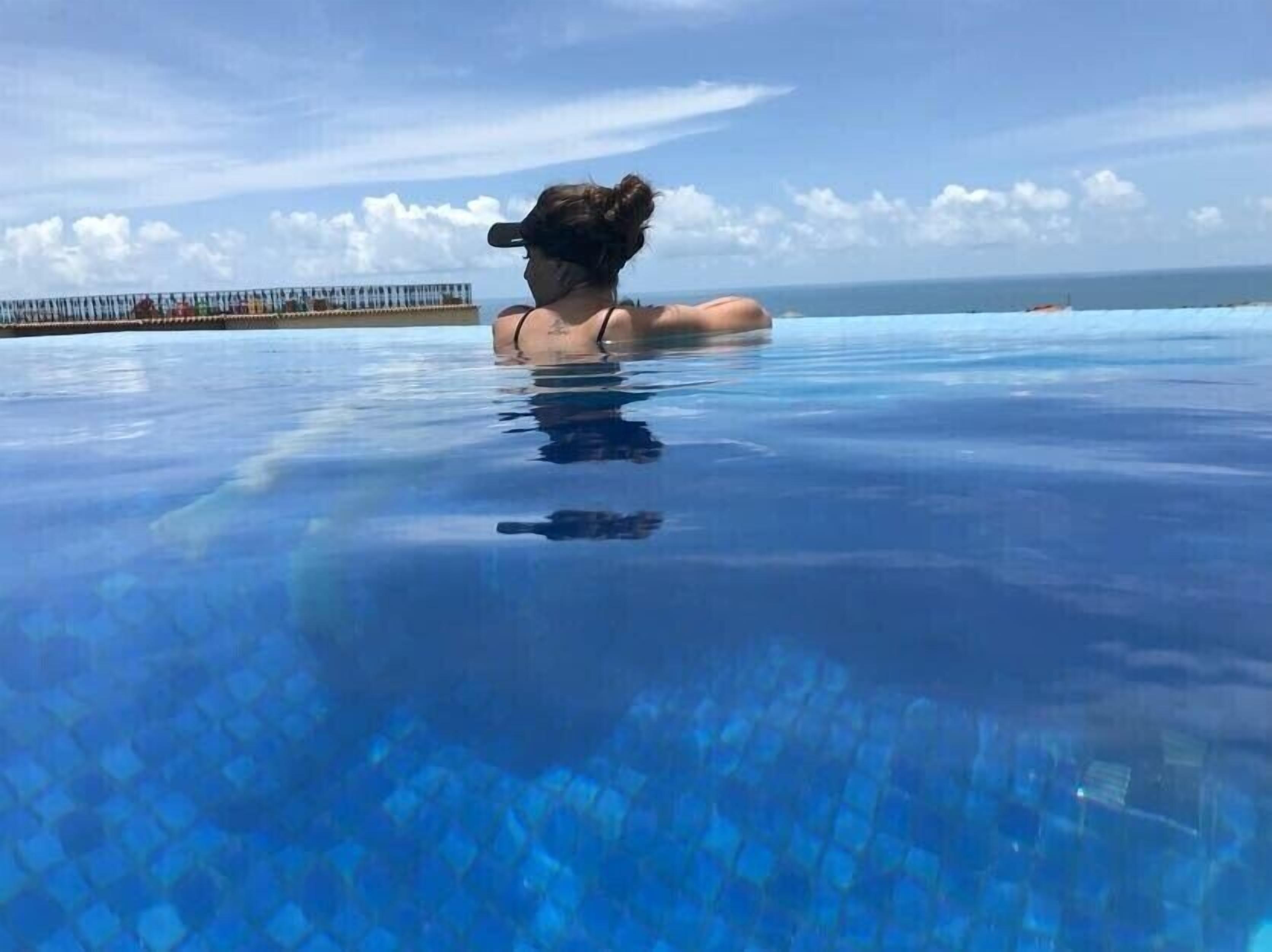En person i en klarblå, oändligt stor pool med utsikt över havet och en avlägsen kustlinje.