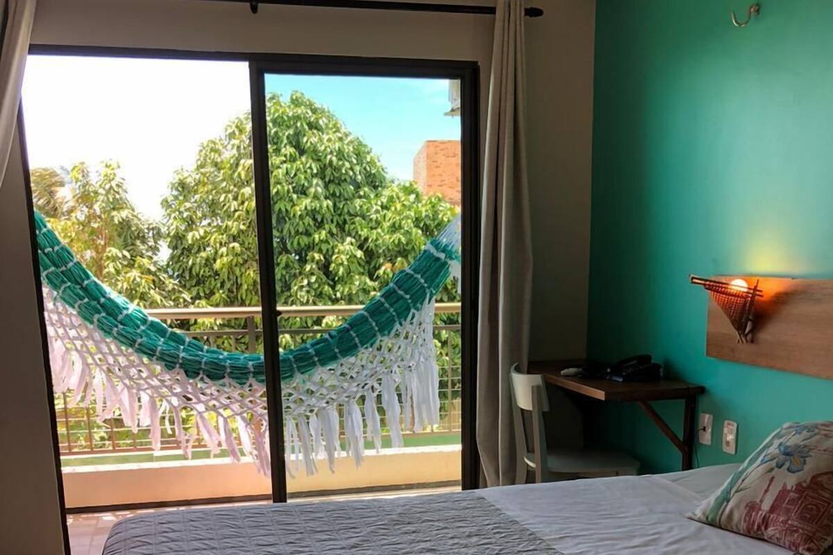 A hotel room with a large bed, a desk, and a view of a green outdoor area with a hammock.