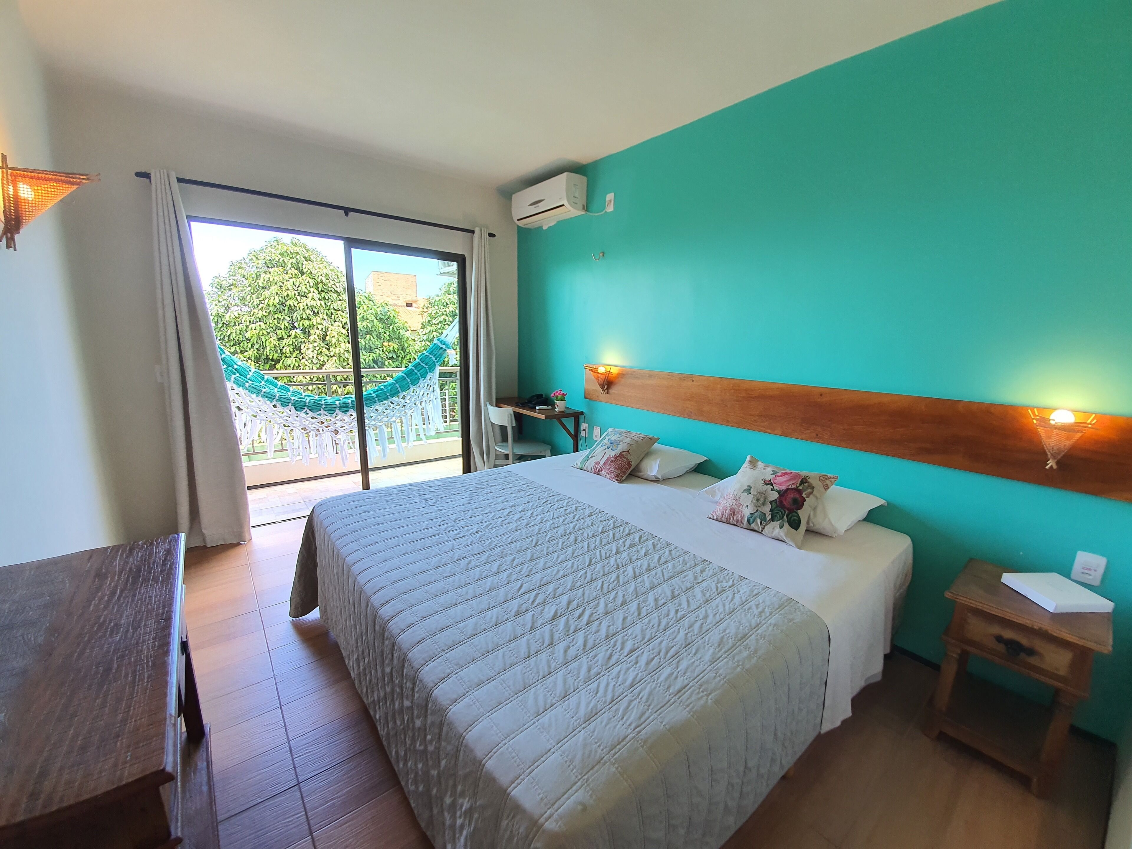Quarto de hotel com cama, mesa de cabeceira de madeira, escrivaninha, cadeira, rede e vista para o verde lá fora.
