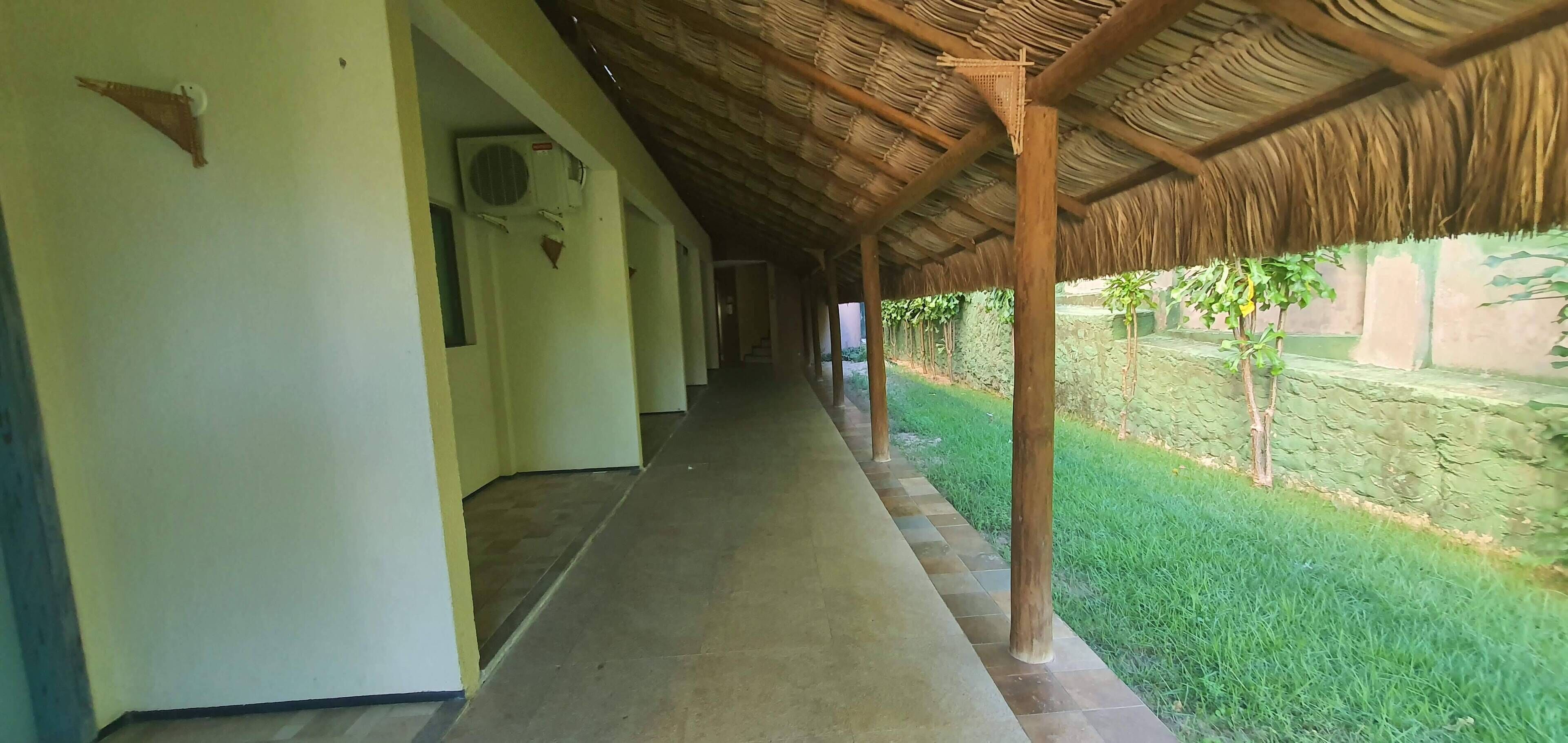 Un corredor cubierto con suelo de baldosas, techo de paja y zona ajardinada con árboles.