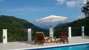 Ein Poolbereich mit zwei Holzliegen, einem weißen Sonnenschirm und Blick auf eine gebirgige Landschaft.