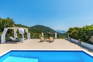 Ein Poolbereich mit Blick auf einen Berg, zwei Liegestühle und eine Pergola mit weißen Vorhängen.