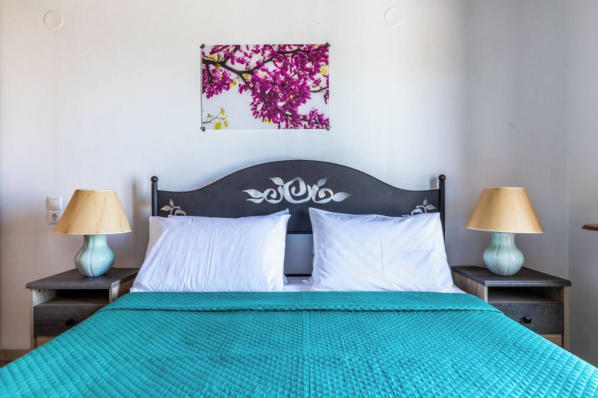 A bed with a headboard, two bedside tables with lamps, a teal bedspread, and a wall-mounted picture of pink flowers.