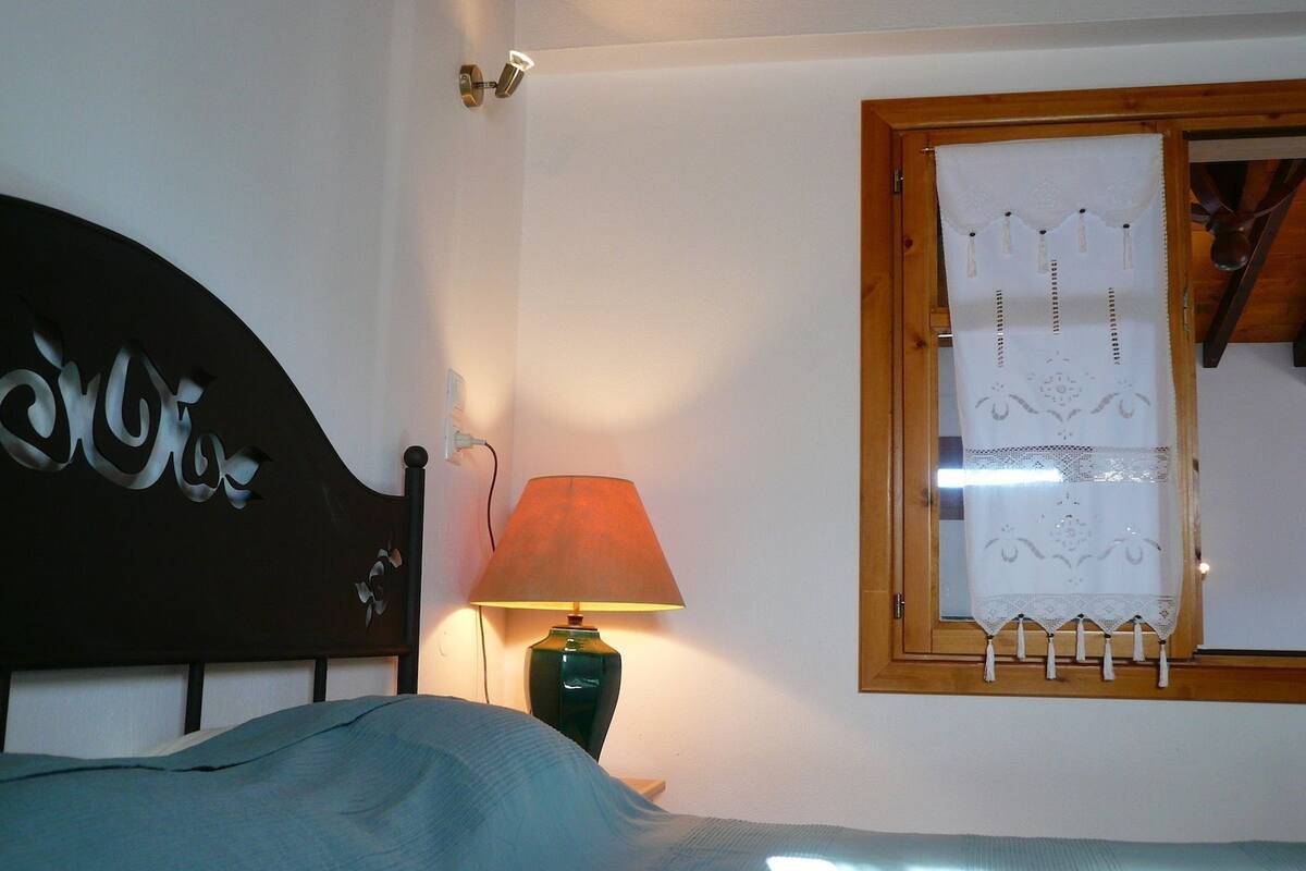 A bedroom with a bed, a lamp, a window with a curtain, and a wall-mounted light fixture.
