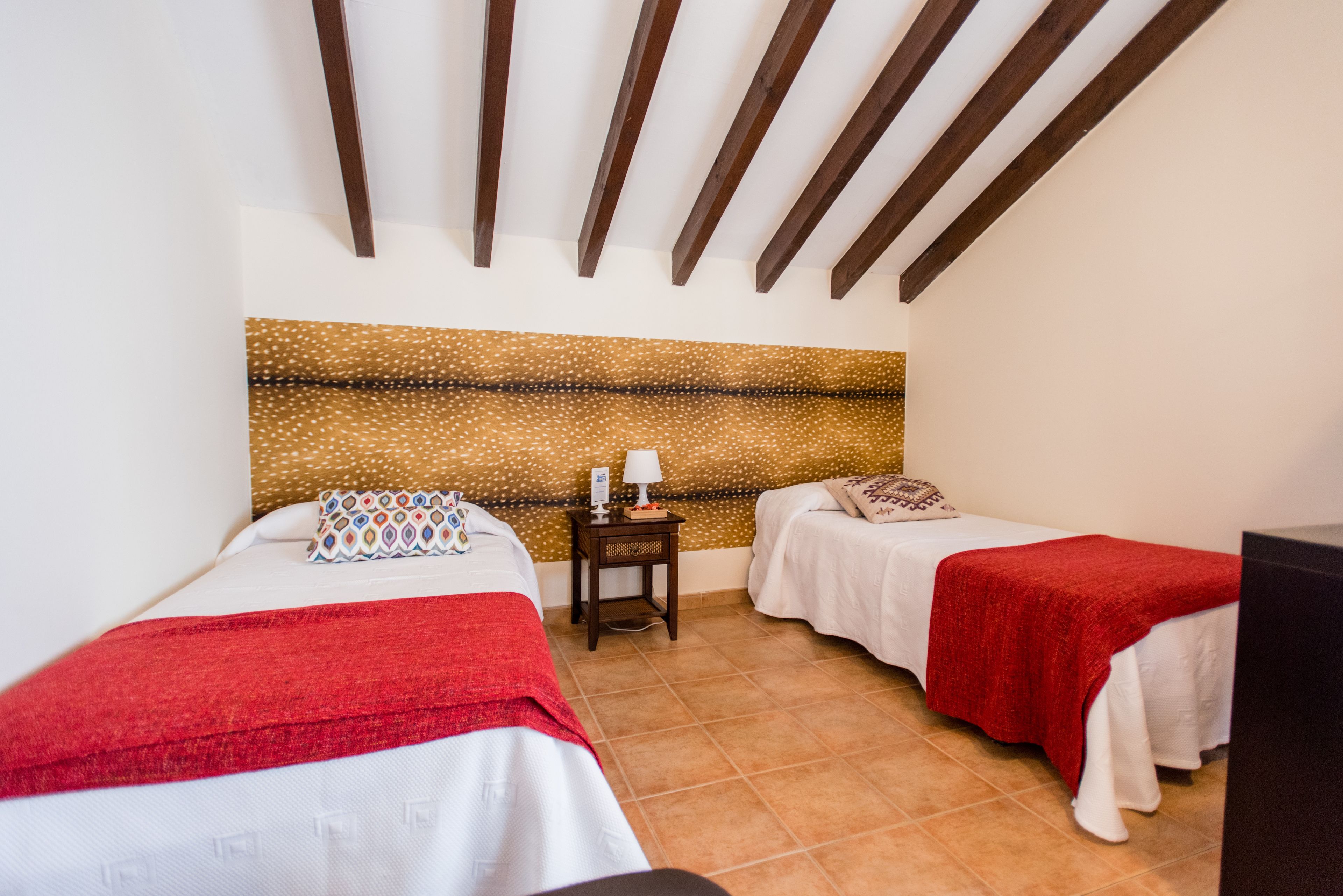 Una habitación con dos camas, cada una con una manta roja, una mesita de noche pequeña con lámpara y un panel decorativo en la pared.