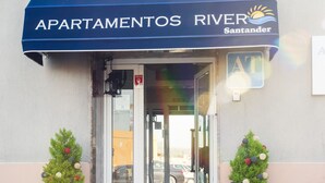 Entrada de un edificio con un toldo azul que dice "APARTAMENTOS RIVER Santander" y dos árboles navideños en macetas.
