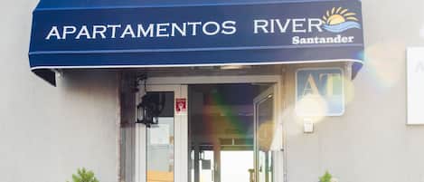 A building entrance with a blue awning labeled "APARTAMENTOS RIVER Santander" and two potted Christmas trees.