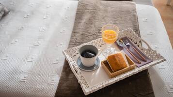 Uma bandeja de café da manhã com xícara de café, copo de suco de laranja, fatia de torrada e talheres sobre uma cama com colcha estampada.