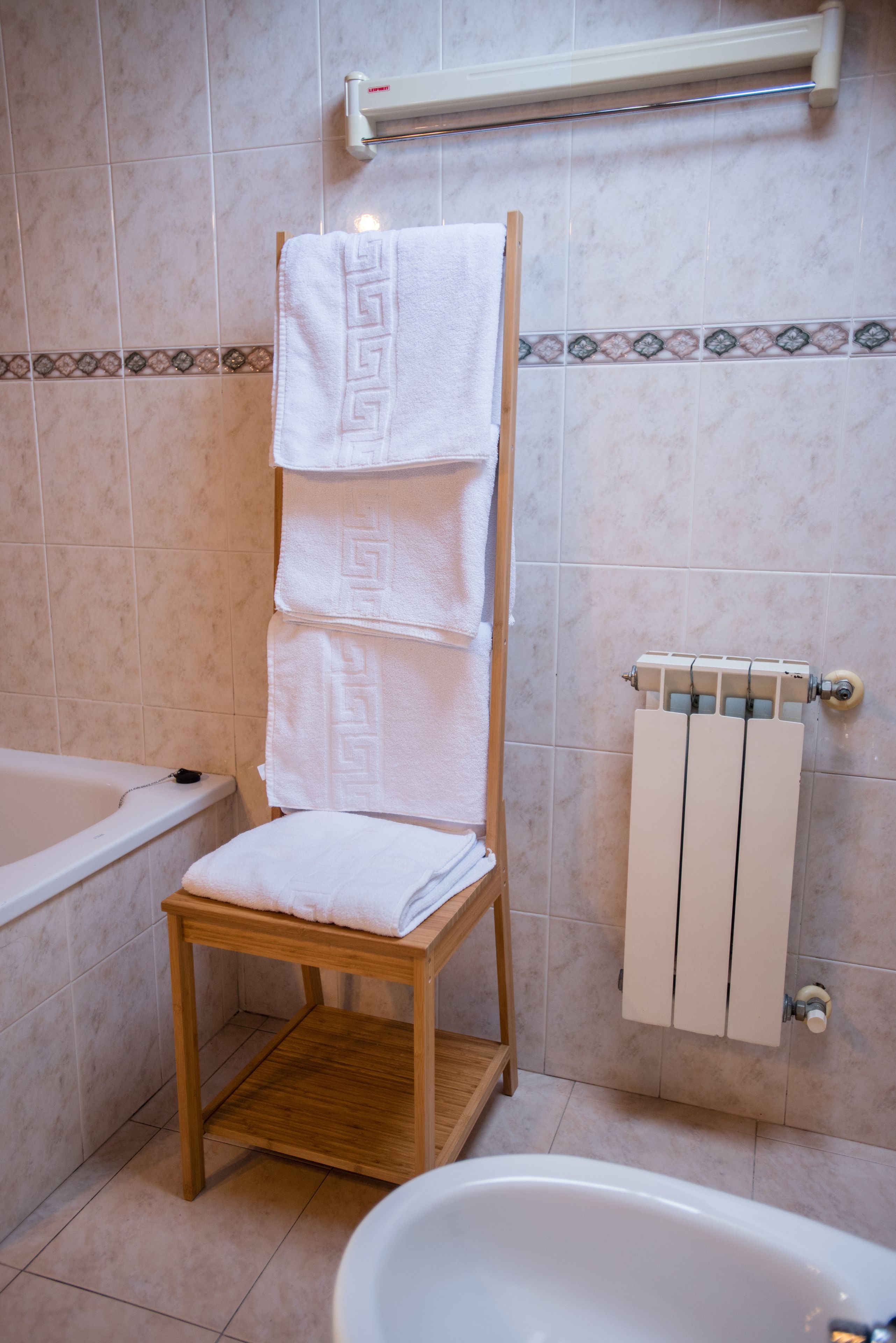 Une salle de bain avec un porte-serviettes en bois sur lequel sont posées trois serviettes, une baignoire blanche, un tabouret en bois et un radiateur blanc.