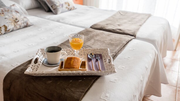Una cama bien tendida con una bandeja de desayuno que incluye una taza de café, un vaso de jugo de naranja y un plato pequeño con un pastel.