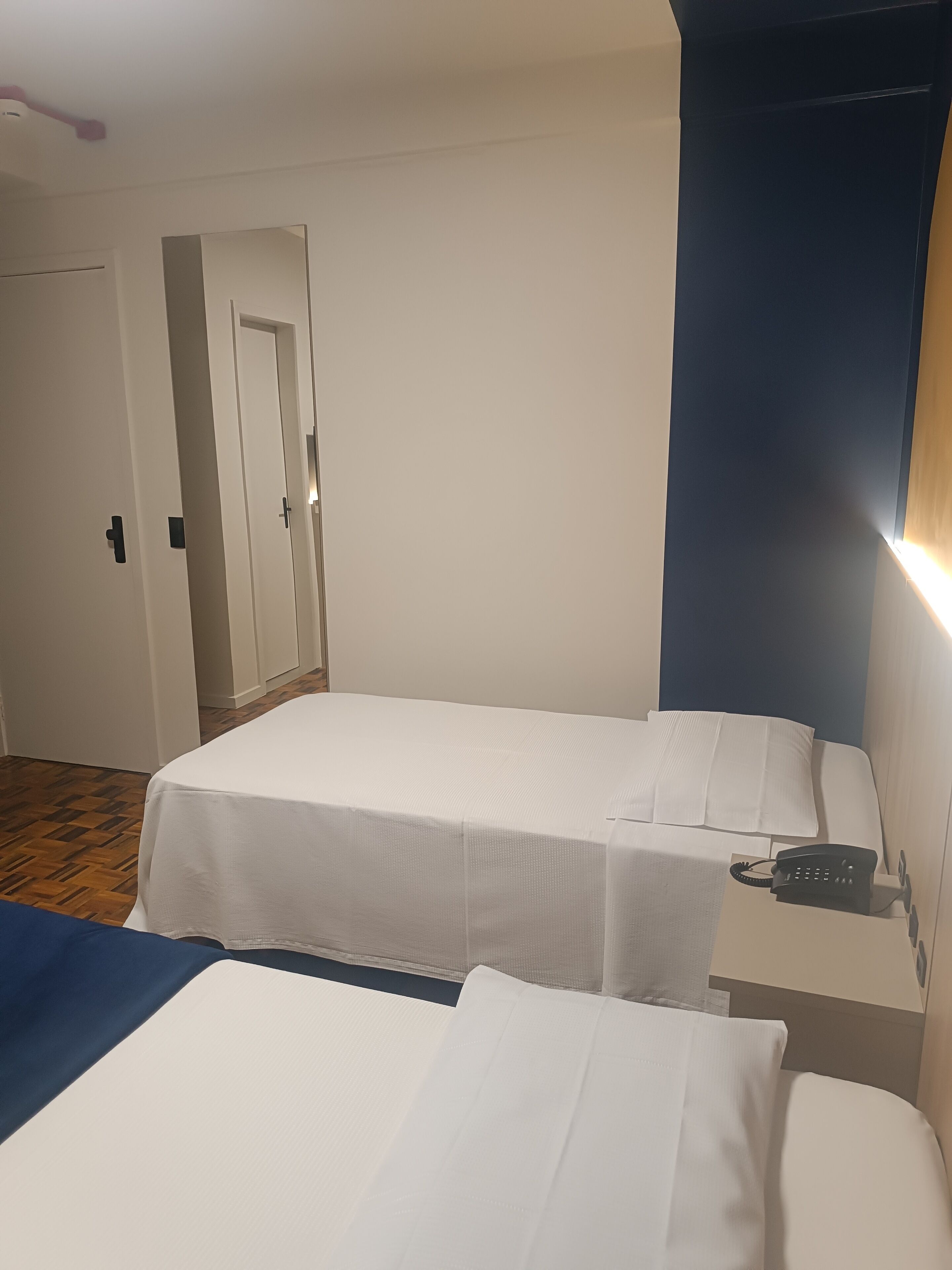 Habitación de hotel con dos camas individuales, un teléfono en la mesita de noche y una puerta que da a otra habitación.