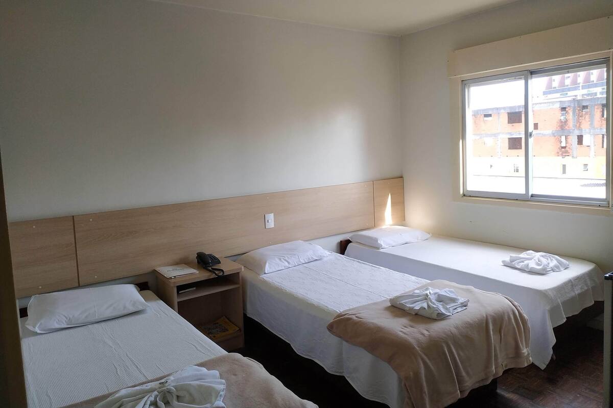Habitación de hotel con dos camas individuales, cabecero de madera, mesita de noche con lámpara y ventana con vistas a edificios.