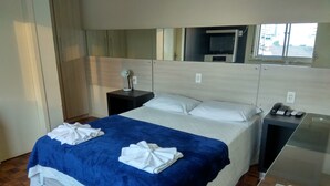 A hotel room with a bed, a bedside table with a phone and remote controls, a mirror, and a window with a view of buildings. - Soratur Hotel & Coworking (Criciuma)