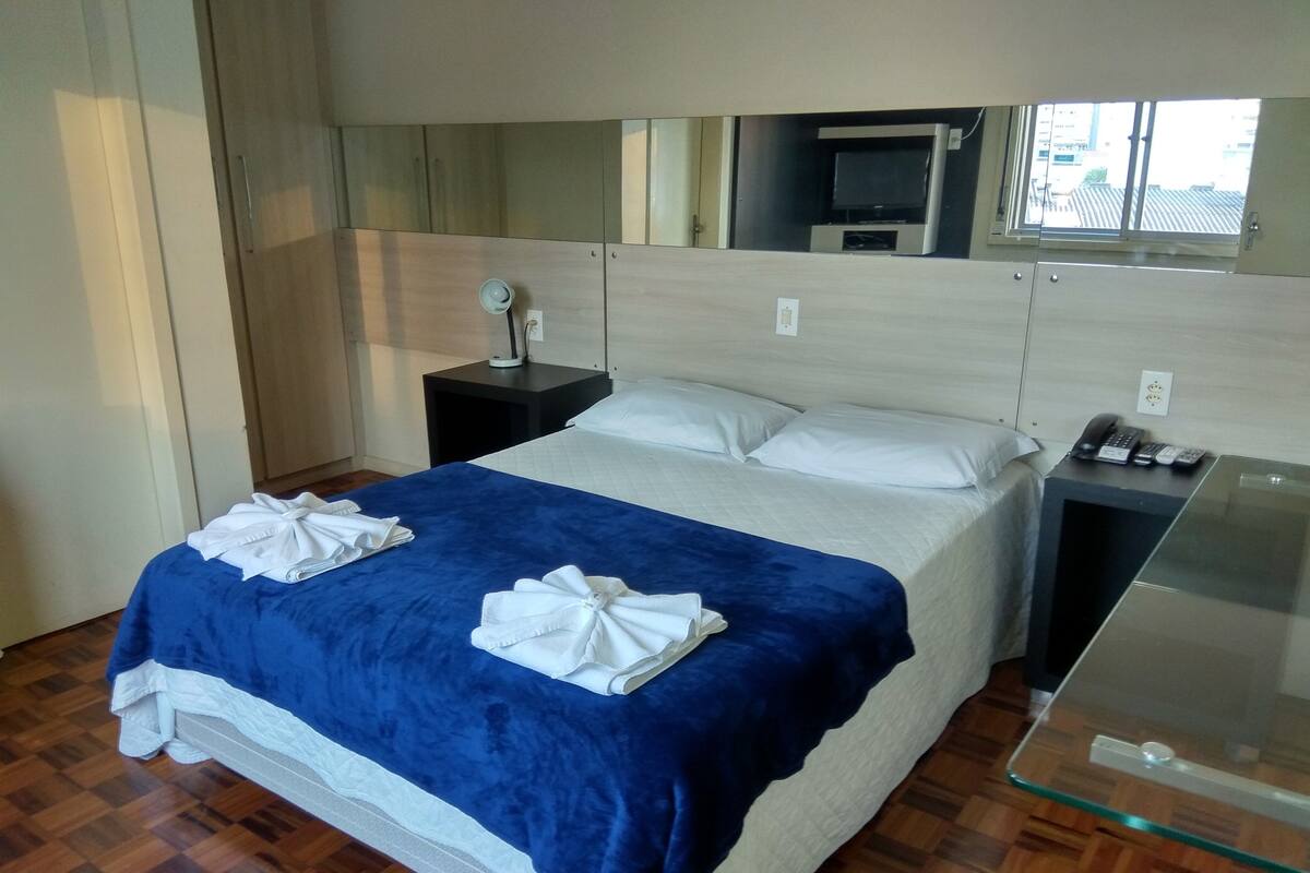 Habitación de hotel con cama, mesita de noche con teléfono y controles remotos, espejo y ventana con vistas a edificios.