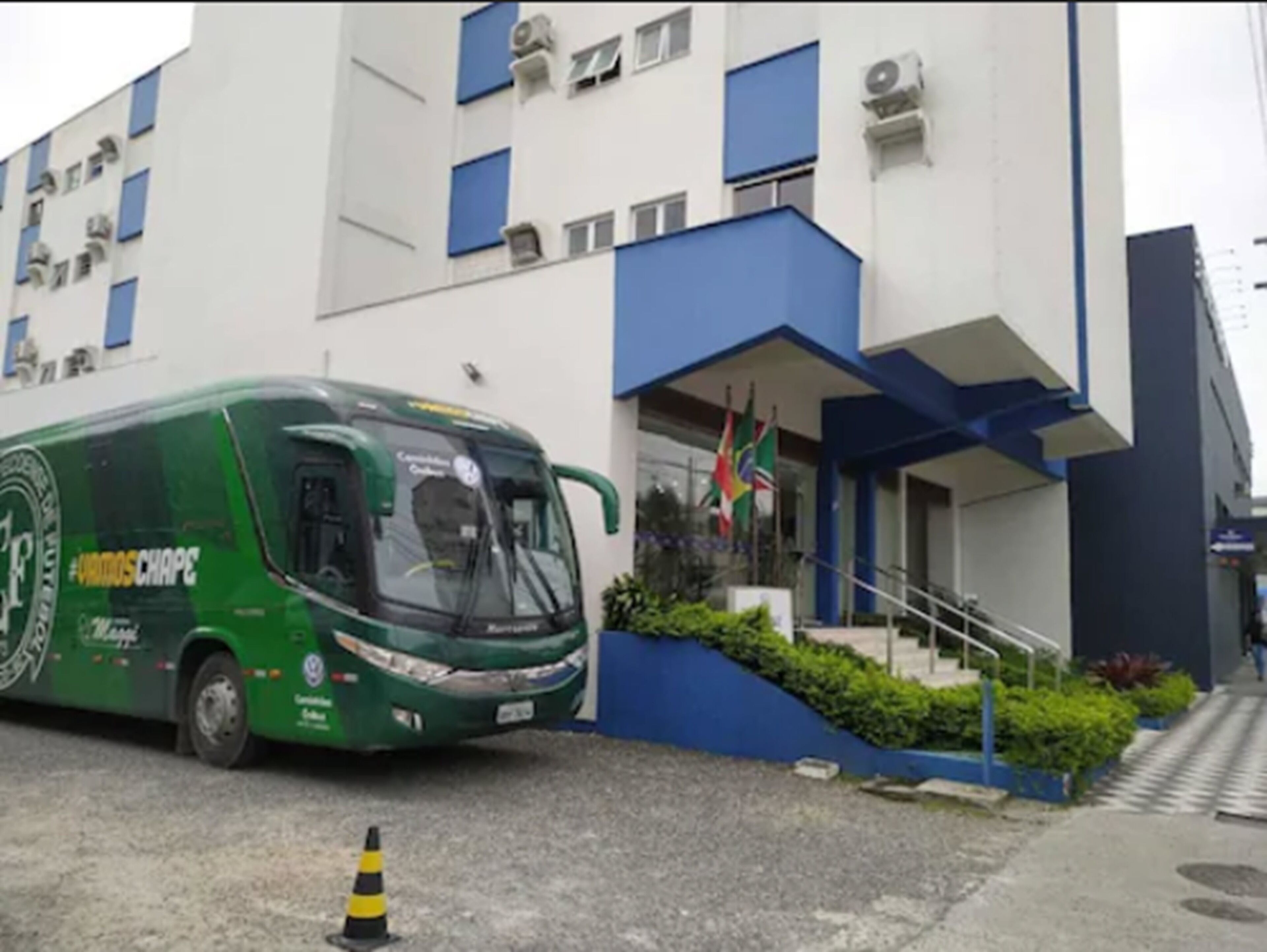Um ônibus verde estacionado em frente a um edifício moderno com detalhes azuis e uma bandeira.
