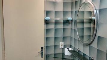 Un baño con pared de baldosas a cuadros, una puerta blanca, repisas de vidrio con artículos de tocador, un espejo redondo y un lavamanos sobre pedestal.