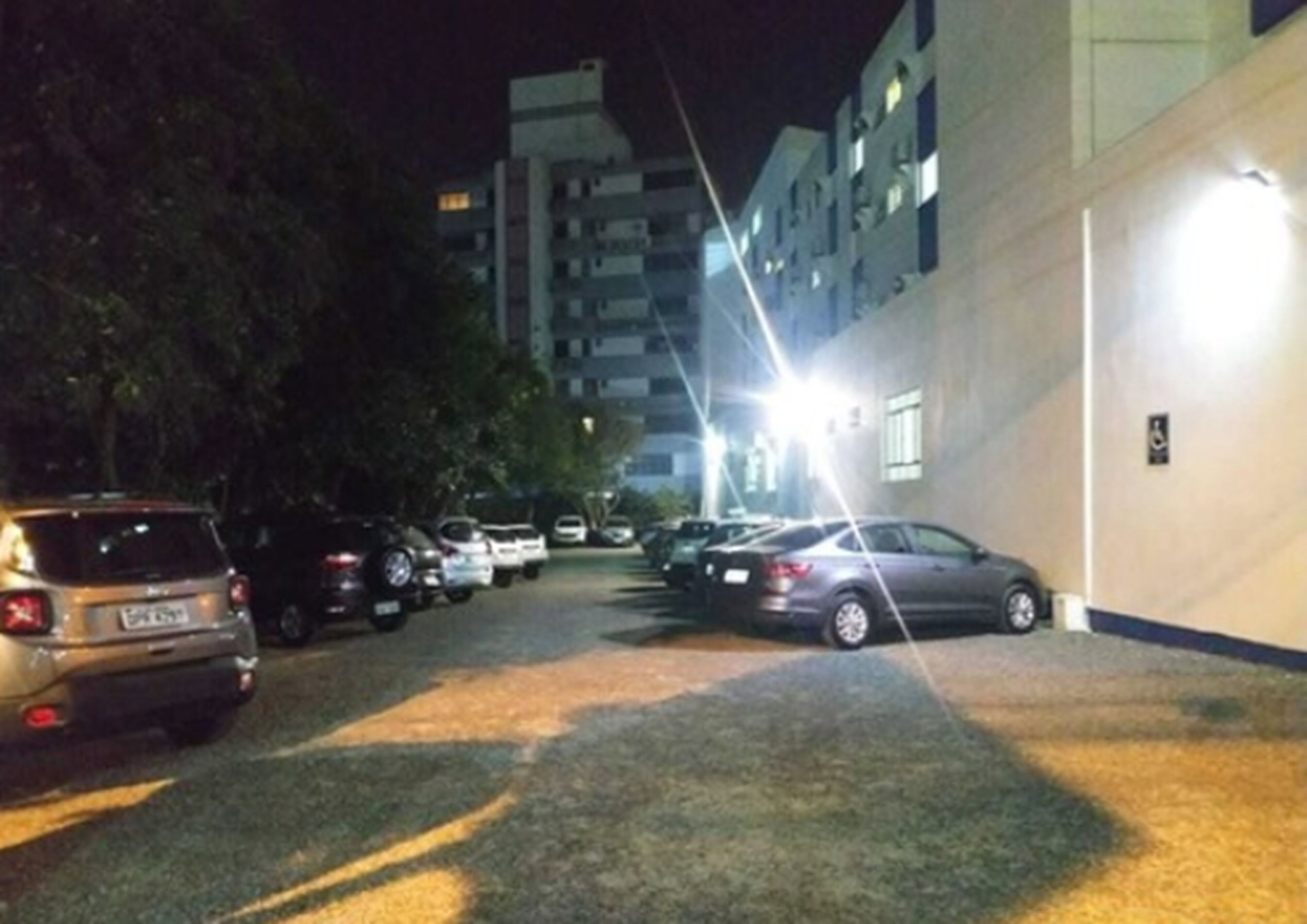 A parking area with multiple cars parked in rows, a building with multiple floors and windows, and a well-lit entrance.