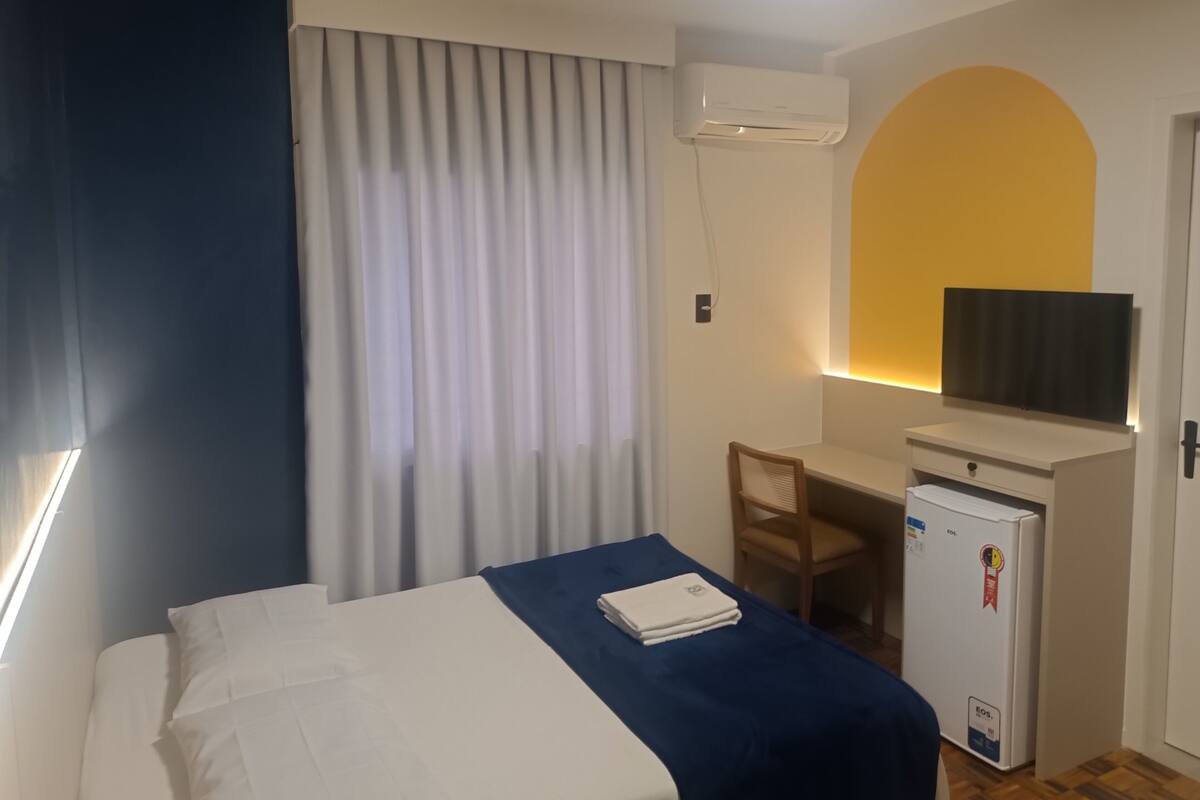 Habitación de hotel con cama, escritorio con silla, refrigerador pequeño y televisor montado en la pared.