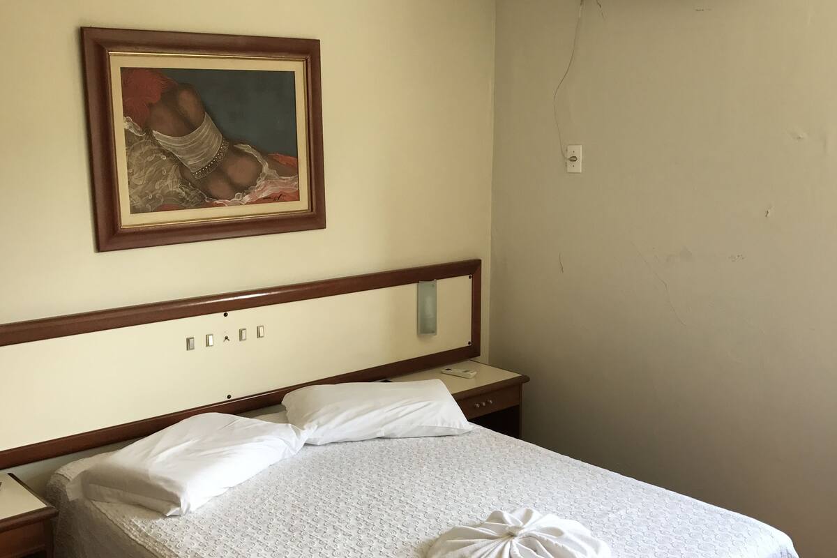 Habitación de hotel con cama, mesita de noche, aire acondicionado instalado en la pared y un cuadro enmarcado sobre la cama.