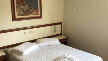 A hotel room with a bed, a bedside table, a nightstand, a wall-mounted air conditioner, and a framed picture above the bed.