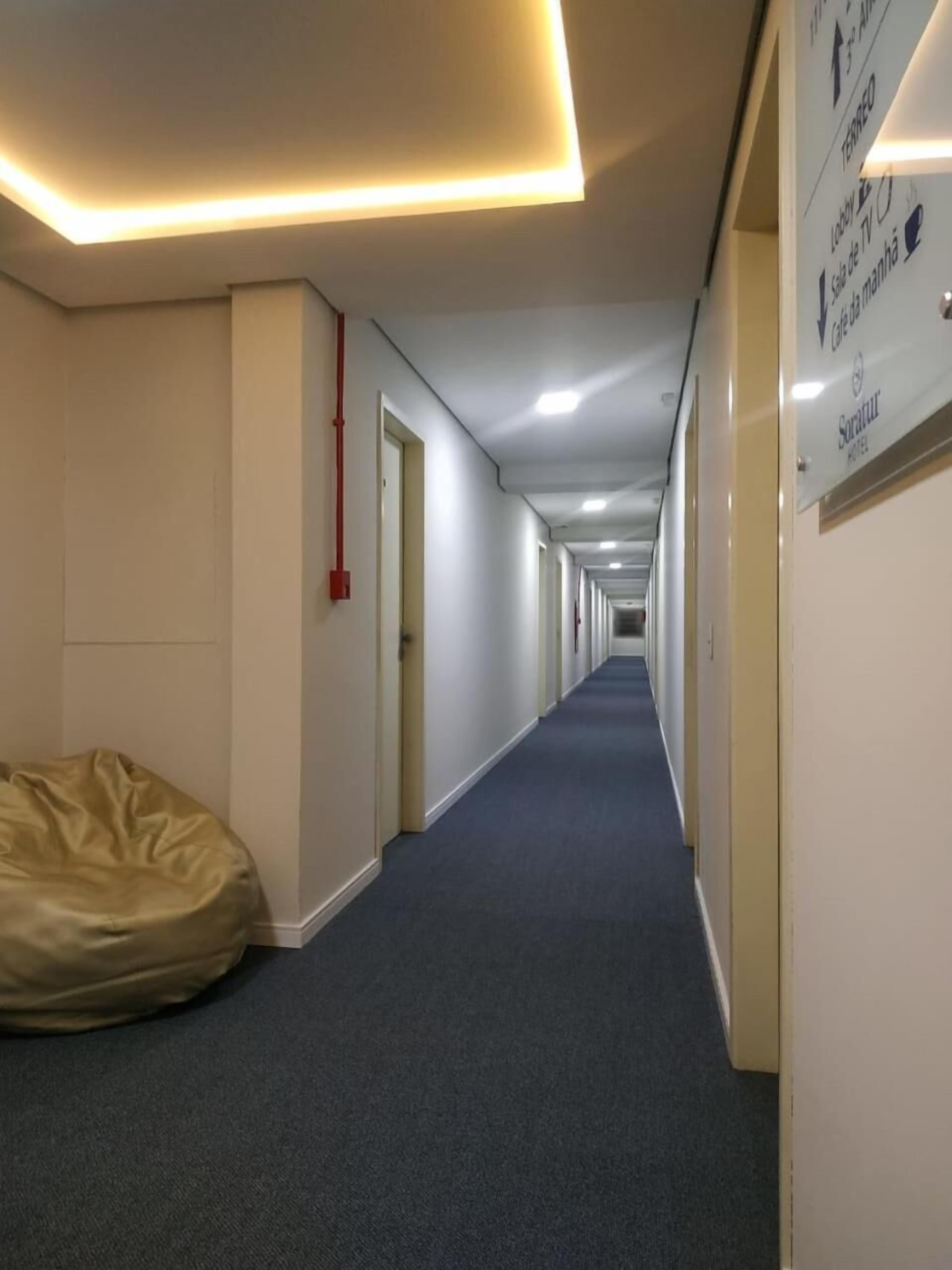 Un pasillo de hotel con diseño largo y estrecho, un sillón informal de color beige y señales direccionales en las paredes.