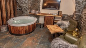 Un cuarto de spa con una bañera de madera, una pared de piedra, un estante de madera con objetos, un sofá de cuero y una pequeña mesa.