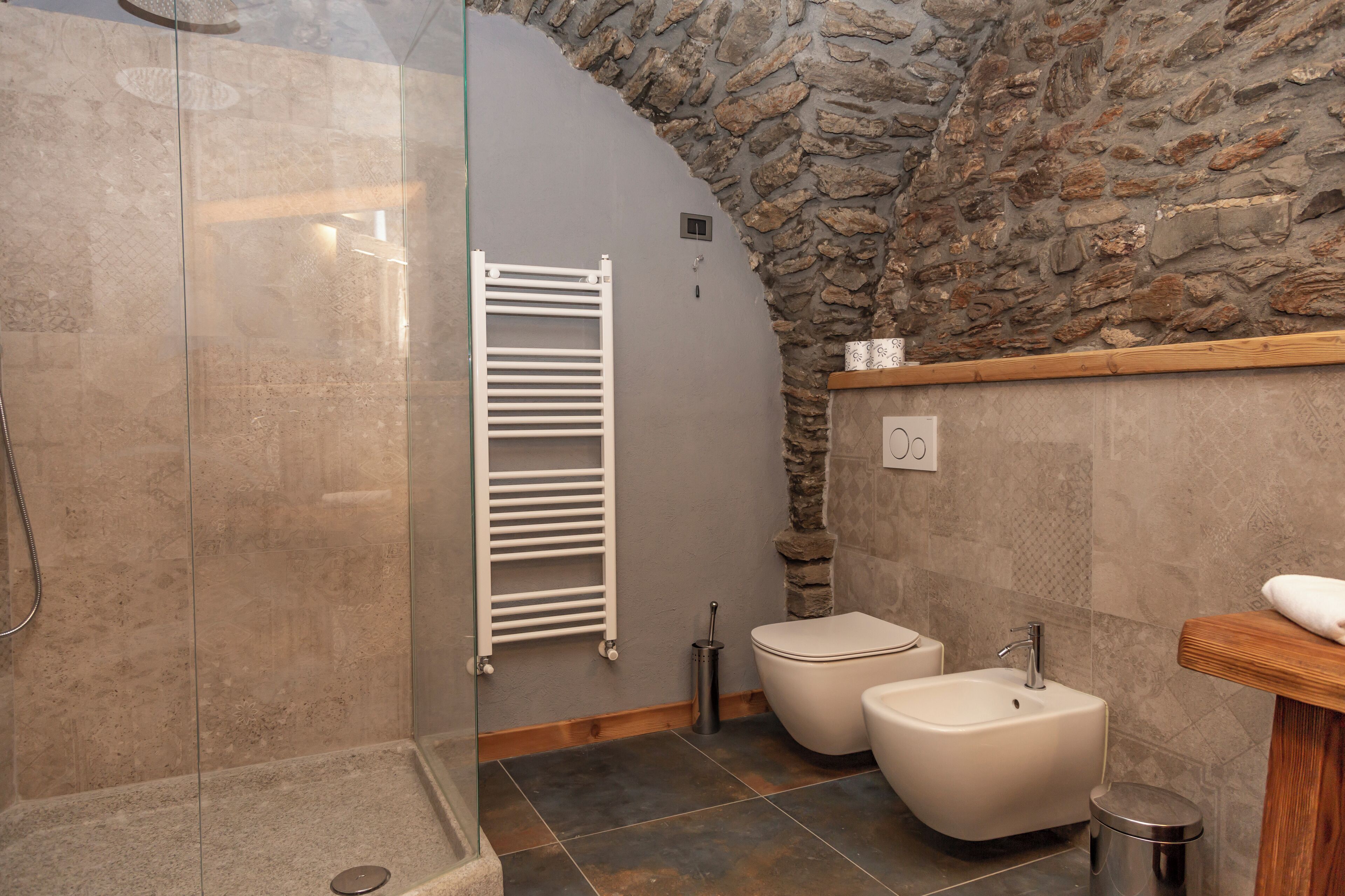 Un baño con cabina de ducha de vidrio, toallero blanco, dos inodoros, un lavabo y una pared de piedra.