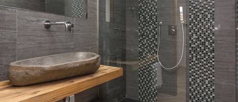Un baño moderno con lavamanos de piedra, estante de madera y cabina de ducha con mosaicos.