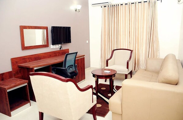 A hotel room with a wooden desk, two chairs, a sofa, a television, and a large window with curtains. - Georgetown Hotel (Abuja)