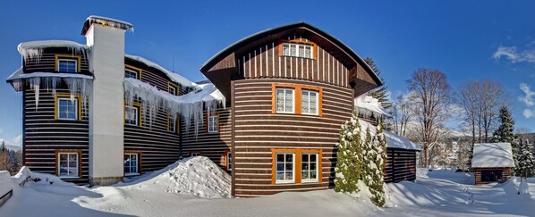 Una cabaña de troncos con una chimenea grande, varias ventanas y un paisaje cubierto de nieve.