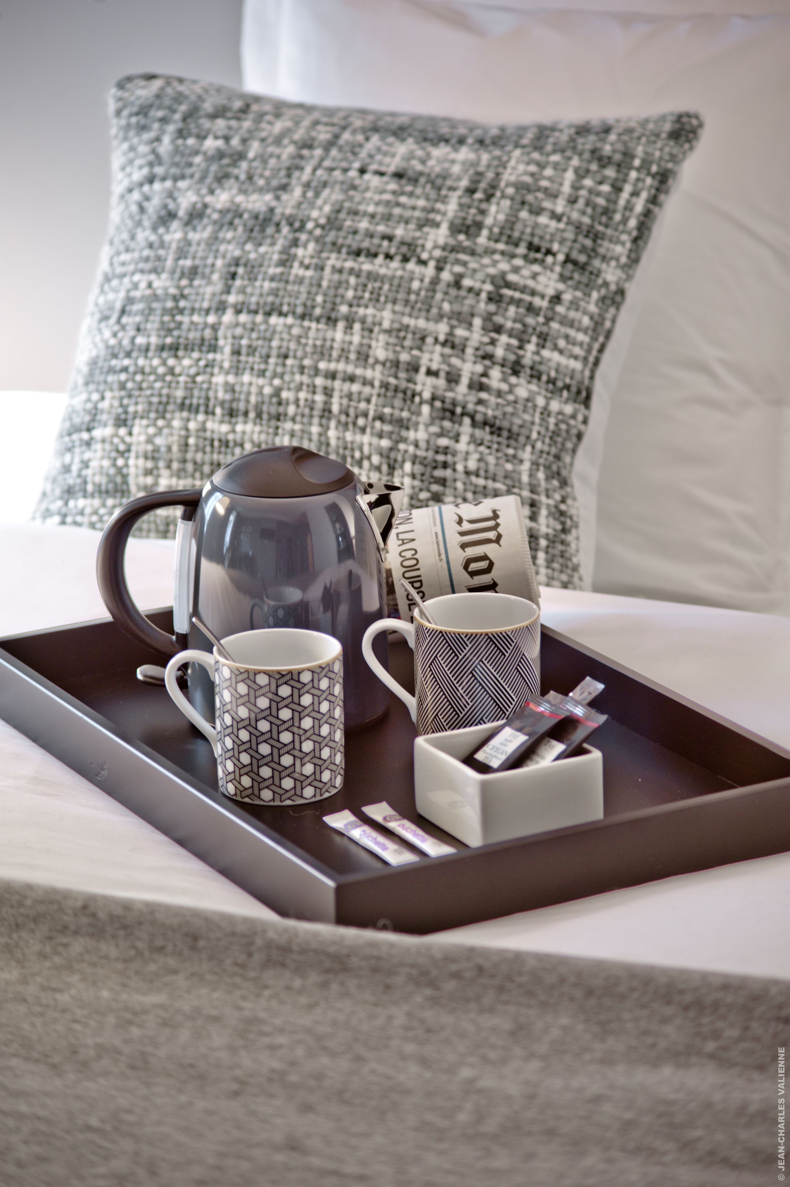 Een dienblad met een koffiepot, twee gemêleerde mokken en een stapel kranten op een bed met een ribbelkussen.