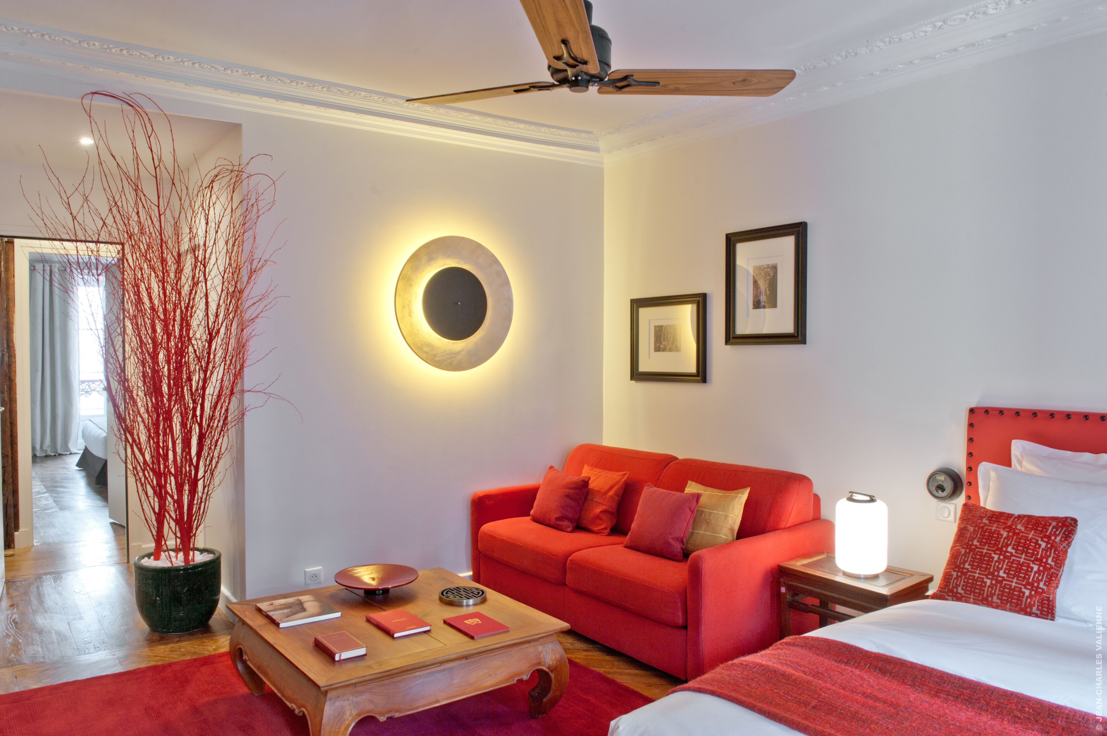 A hotel room with a red sofa, a wooden coffee table, a bed with a red headboard, a ceiling fan, and a wall-mounted circular light fixture.