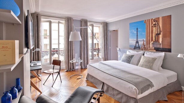 A hotel room with a large bed, a desk with a chair, a small table with a lamp, and a balcony with a view of the Eiffel Tower. - My Home For You B&B (Paris)