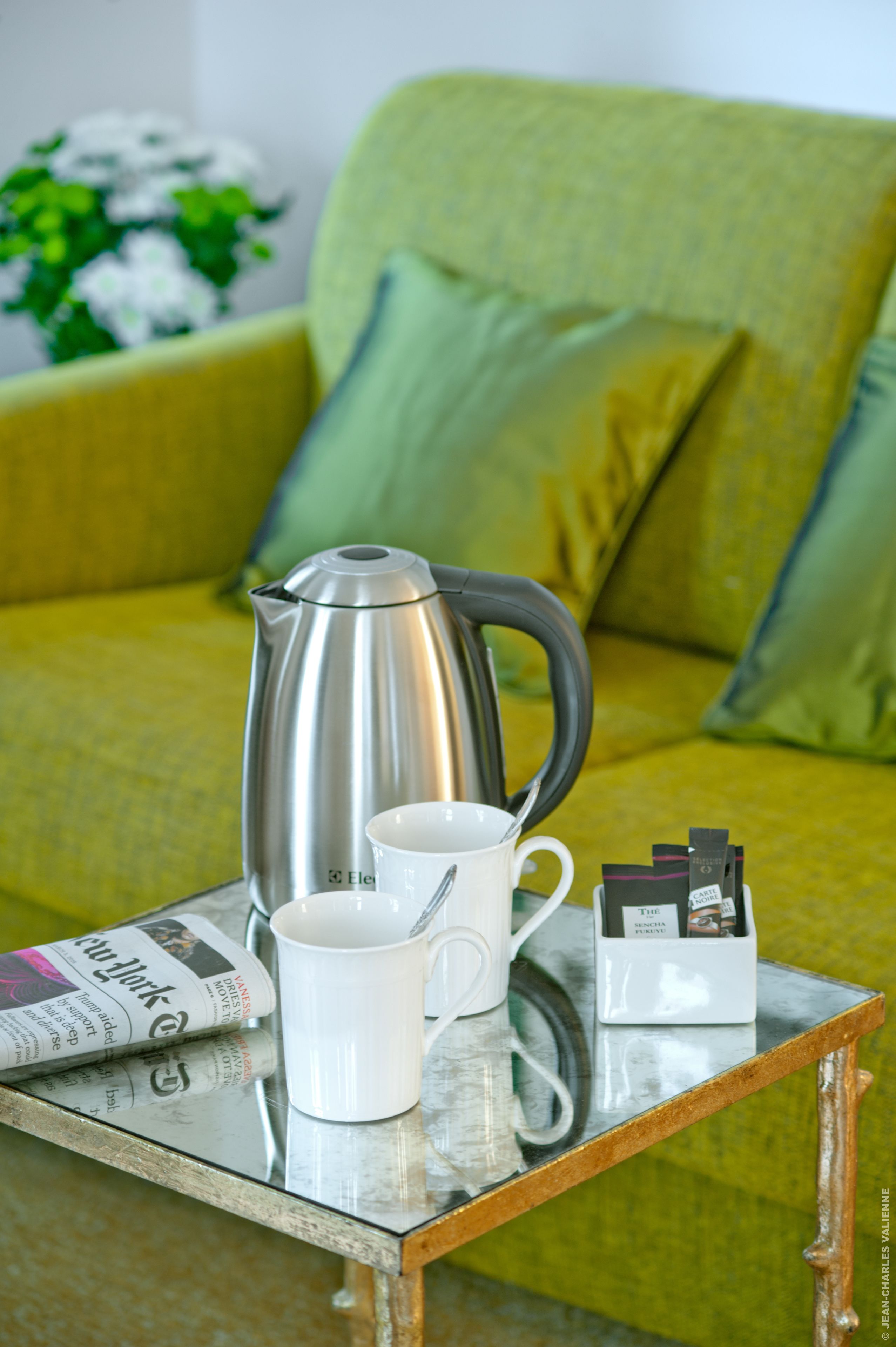 Una mesa de centro con un periódico, una taza blanca, una cafetera de plata y una pequeña bandeja con cápsulas de café.