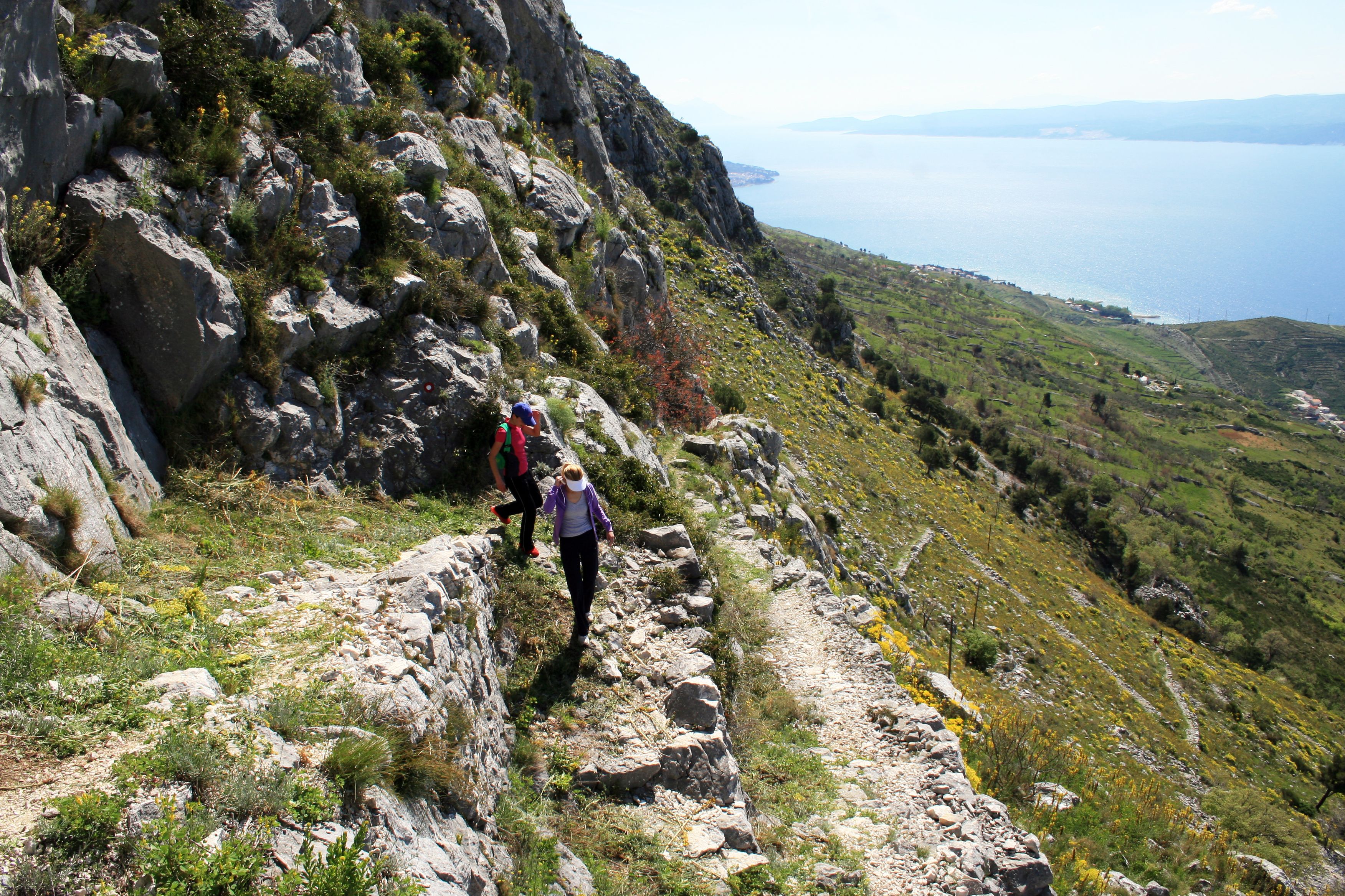 A rocky hiking trail with a view of the sea and distant mountains.
