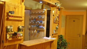 A wooden reception desk with a plant to the right, surrounded by shelves with various items and a door in the background. - Hotel Výsluní (Spindleruv Mlyn)