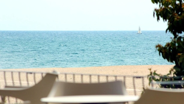 Vista de una playa con un velero a lo lejos, una orilla arenosa y mobiliario exterior.