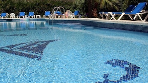 Piscina cristalina con mosaicos azules, rodeada de sillas de descanso azules y una palmera.