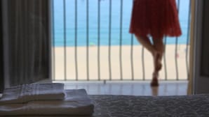 Una habitación con cama, ventana y vista a la playa, y una persona con un vestido rojo.