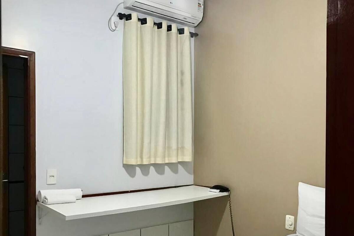 Habitación de hotel con cama, inodoro blanco, toalla, espejo, ventana con cortinas y unidad de aire acondicionado.