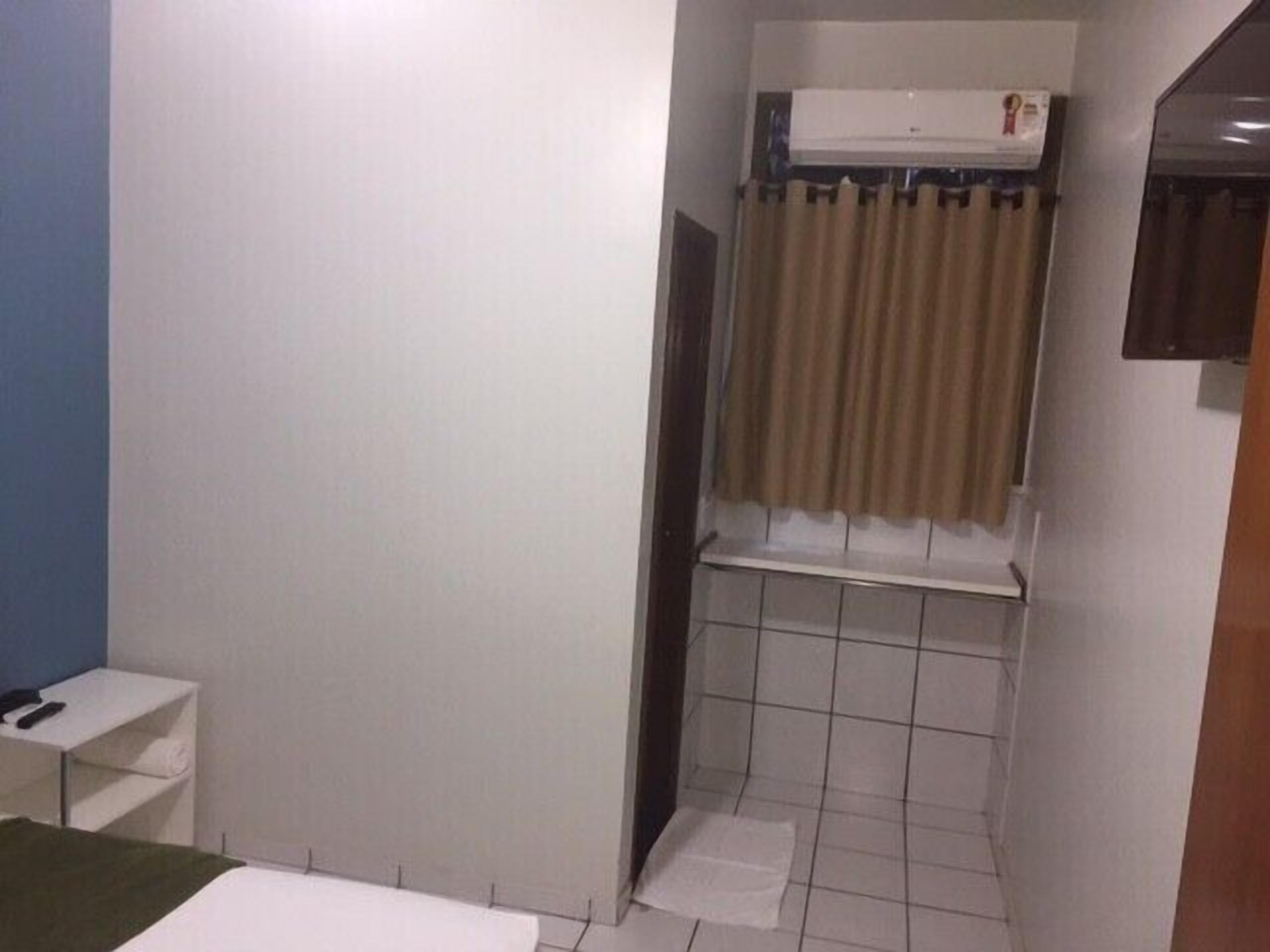 A small, compact room with a single bed, a white bedside table, a small shelf, a window with curtains, and a tiled shower area.