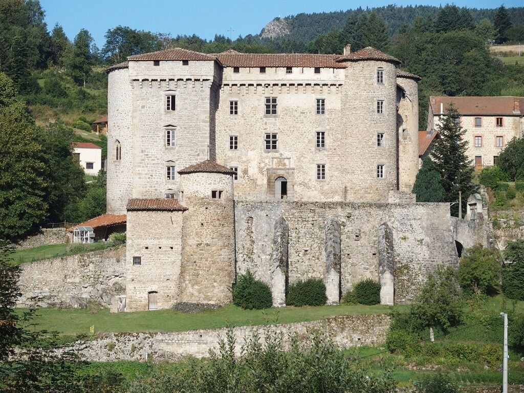 A historic stone building with multiple towers and a fortified wall, surrounded by greenery and other buildings.