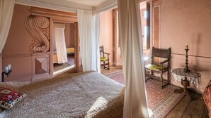 Un dormitorio con una cama grande, una silla, una mesa con una vela y vistas a otra habitación por una puerta abierta.